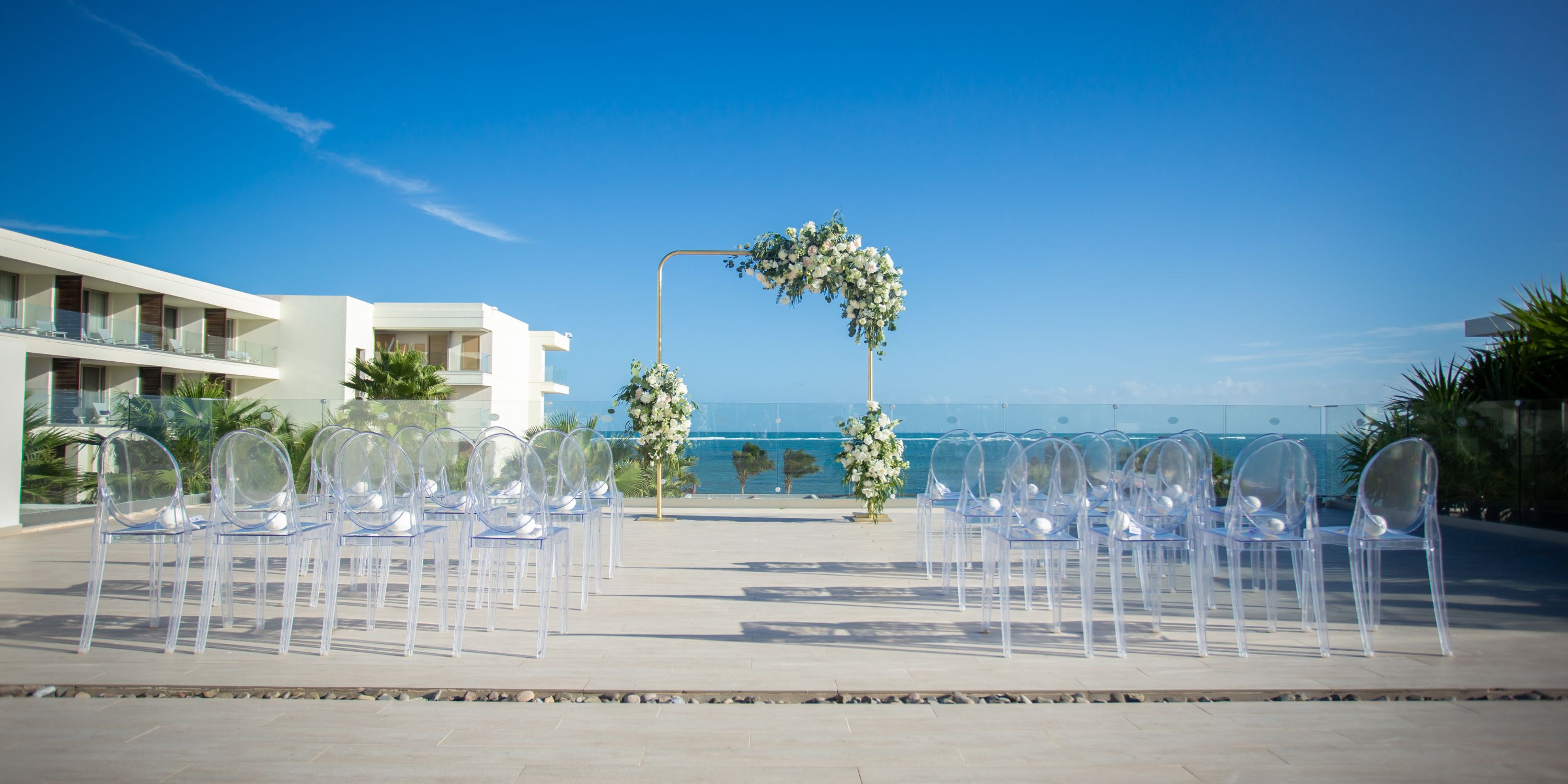 Destination wedding setup at Secrets Riviera Cancun with clear chairs and a floral arch by the ocean.