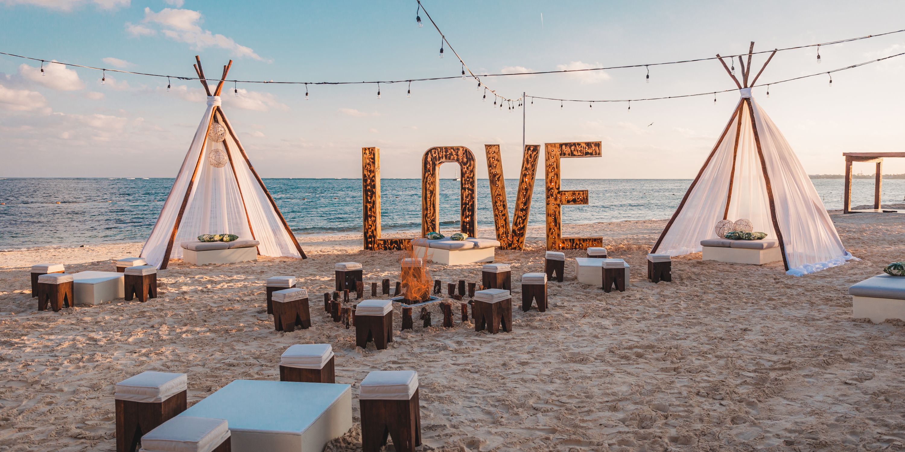 Destination wedding setup at Secrets Riviera Cancun with "LOVE" sign, tipi tents, string lights, and oceanfront seating.