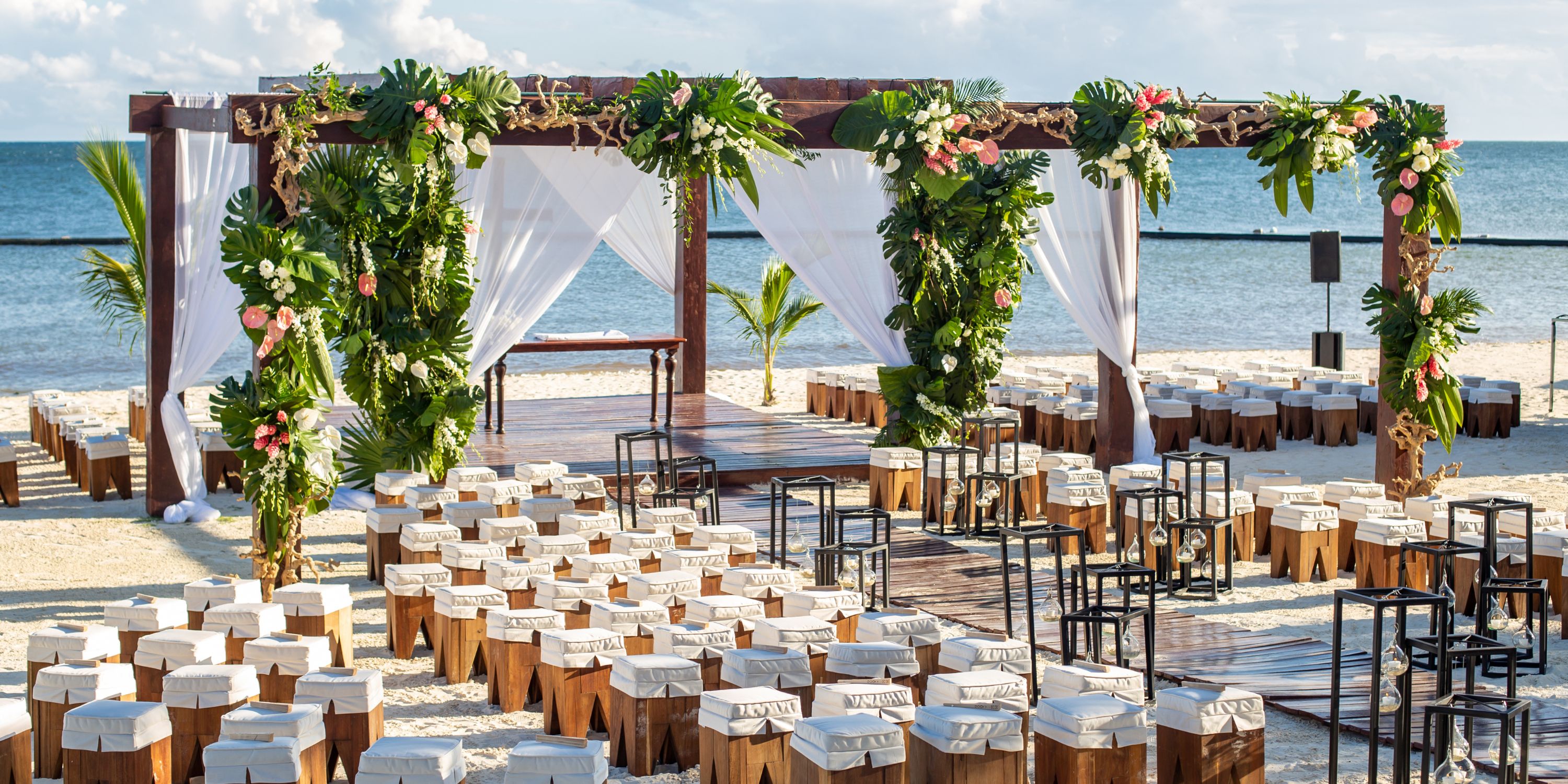 Destination wedding at Secrets Riviera Cancun with wooden seating, floral arch, and ocean in the background.
