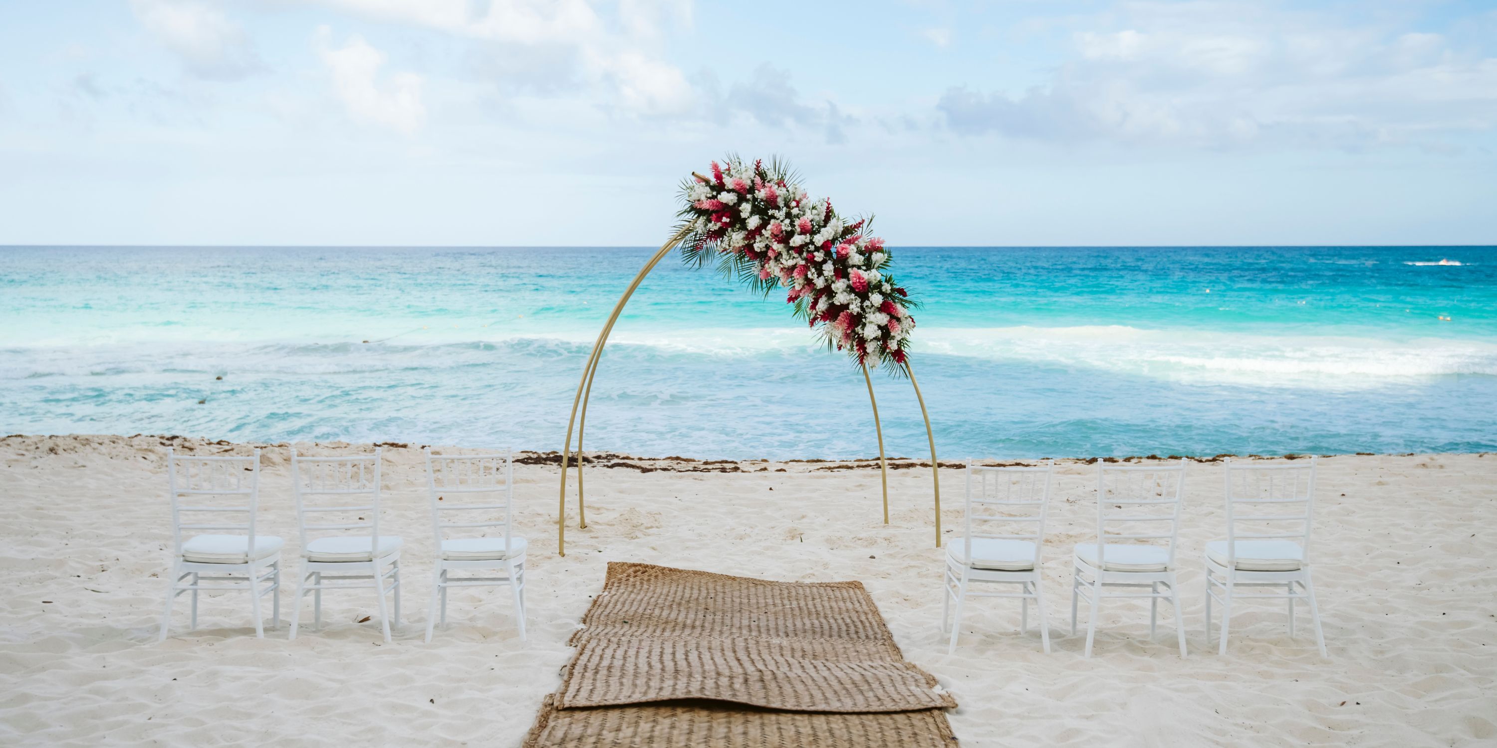 Playacar Palace destination wedding: floral arch on the beach, white chairs, and a woven mat aisle.