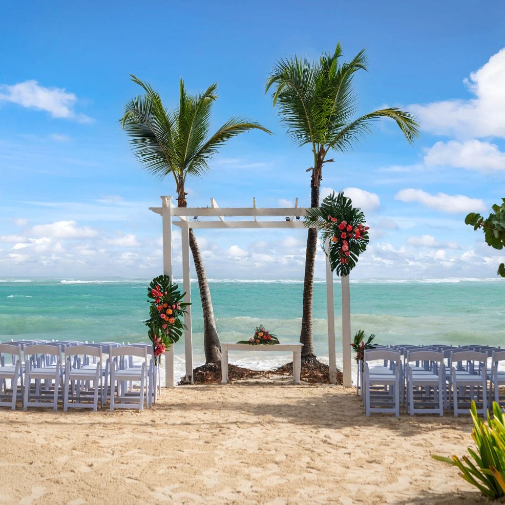 Destination wedding setup on the beach with white chairs, floral arch, and palm trees overlooking the ocean.