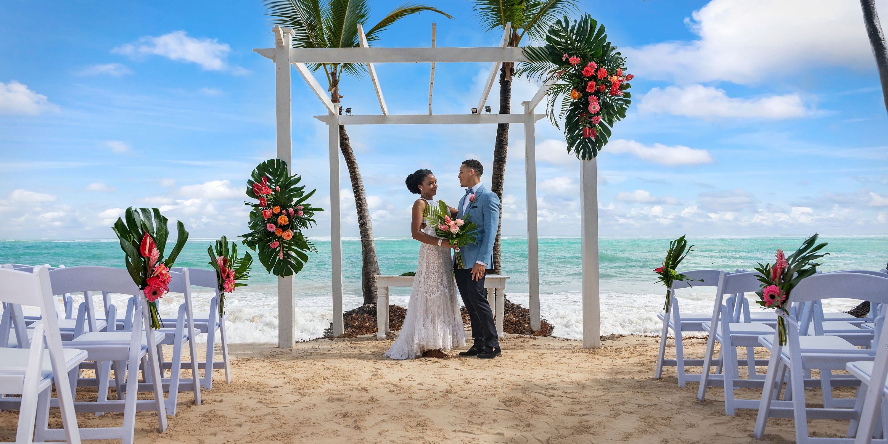 A couple stands under a floral arch at TRS Turquesa during their destination wedding, chairs decorated.