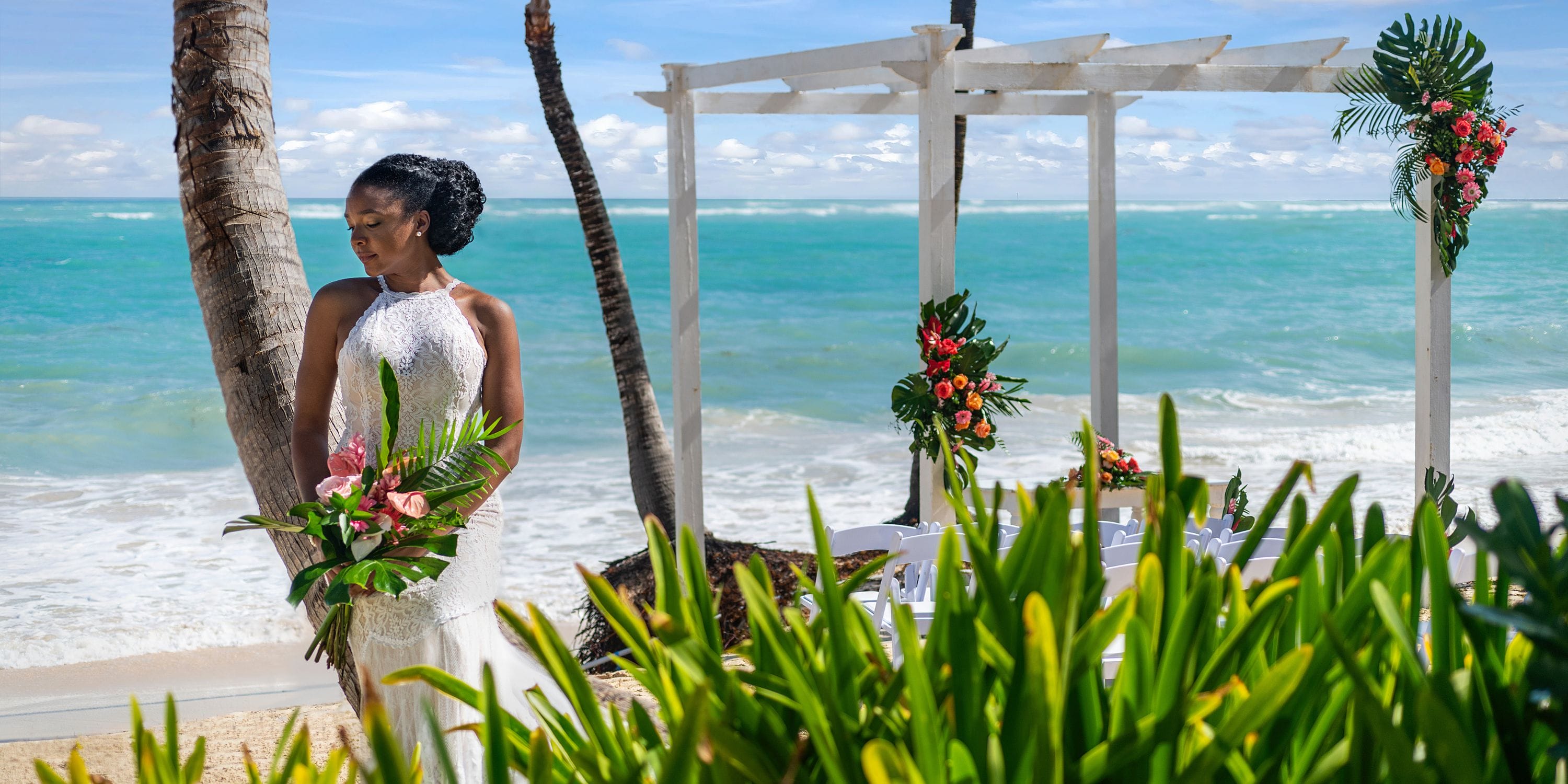 Bride with bouquet stands beside TRS Turquesa wedding arch at a tropical destination wedding beach.