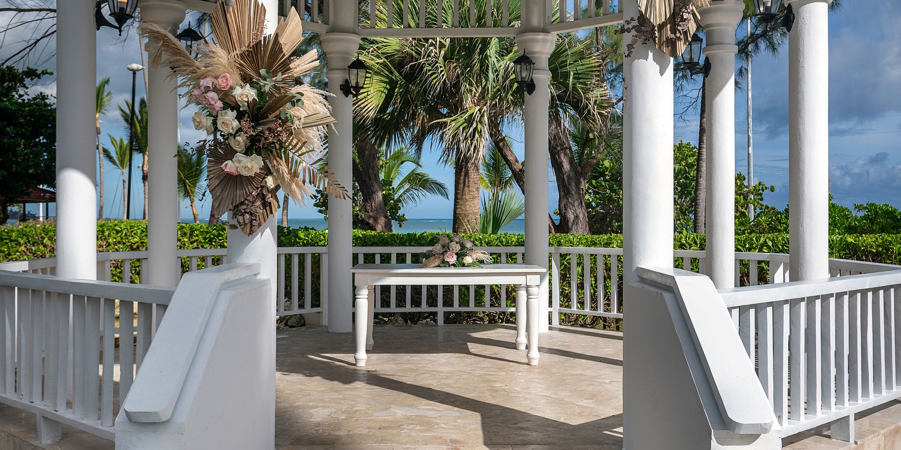 TRS Turquesa: White gazebo with wedding floral decor, nestled in lush greenery and palm trees.