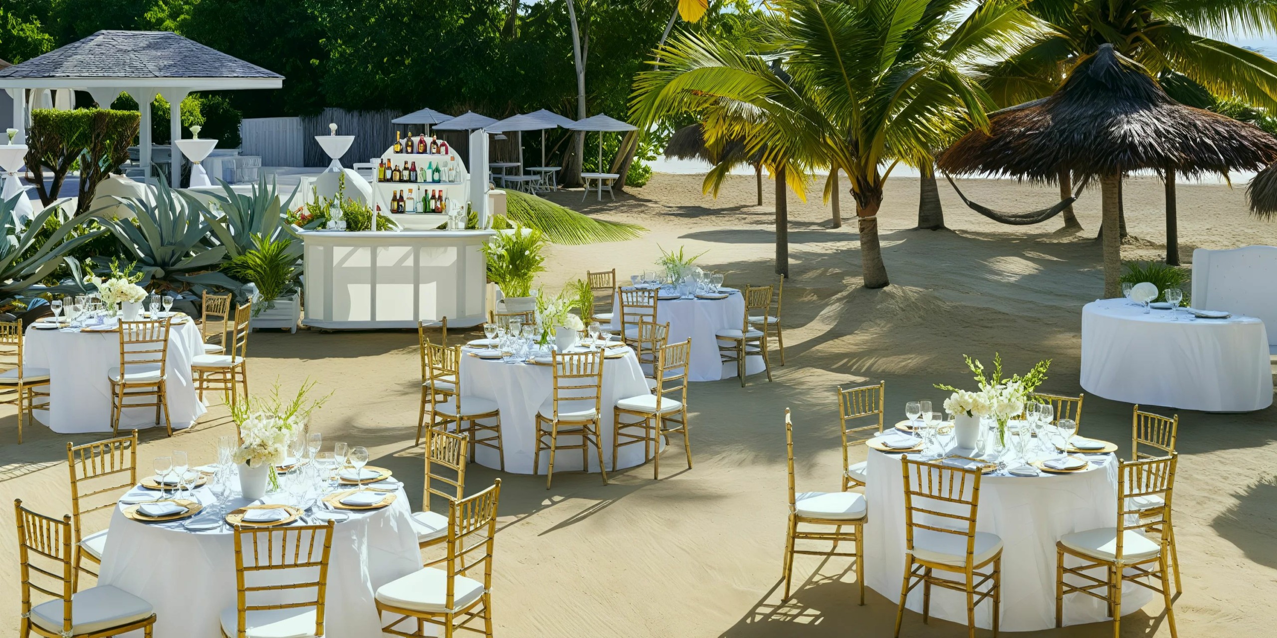 Outdoor beach wedding setup with round tables and white tablecloths, gold chairs, and a bar. Palm trees and white floral centerpieces complete the elegant scene.