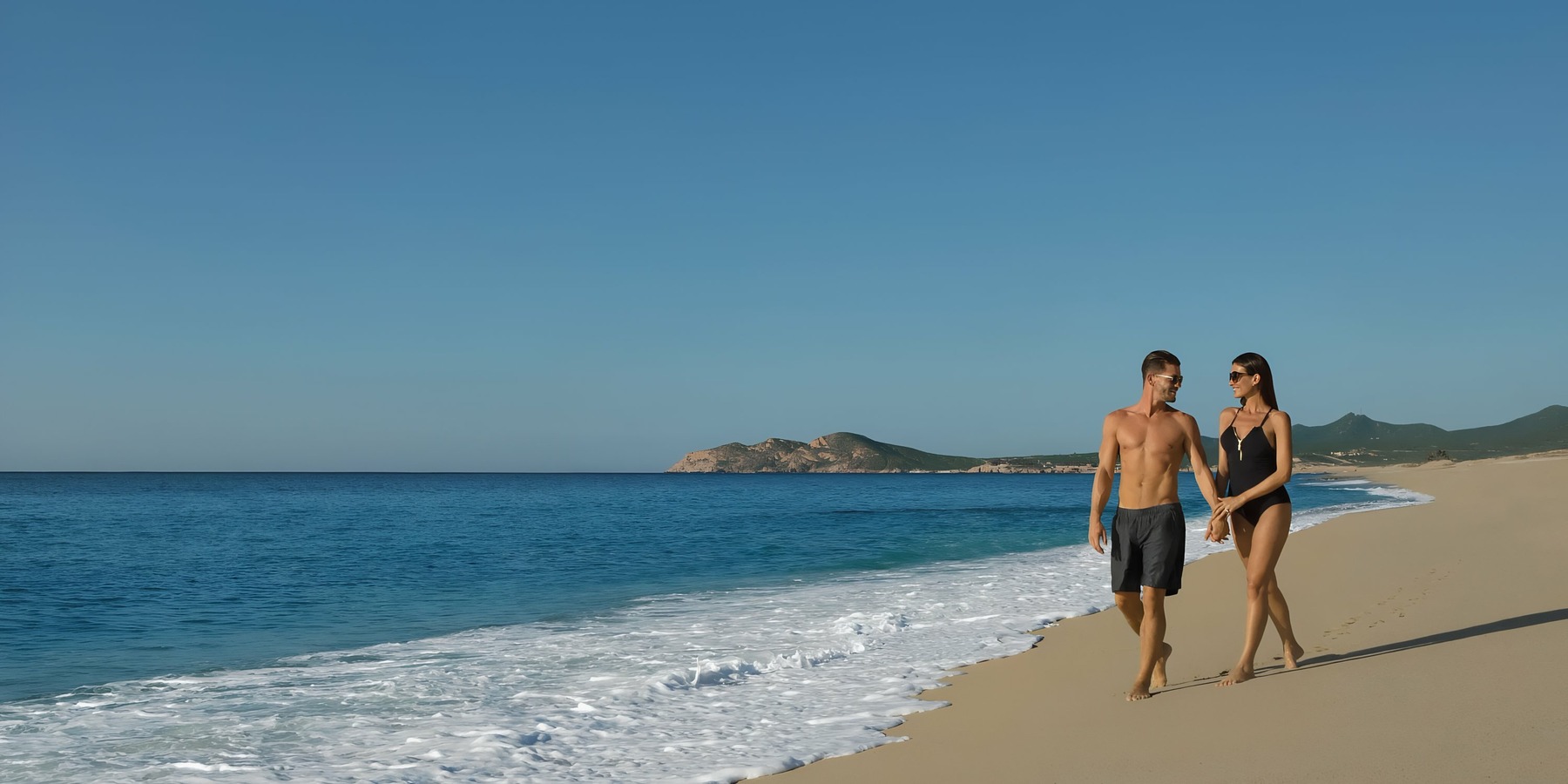 A couple walking along a serene beach with clear skies at Dreams Los Cabos.