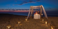 At Dreams Los Cabos, a couple sits at a candlelit table under a canopy on the beach at sunset, surrounded by lanterns.
