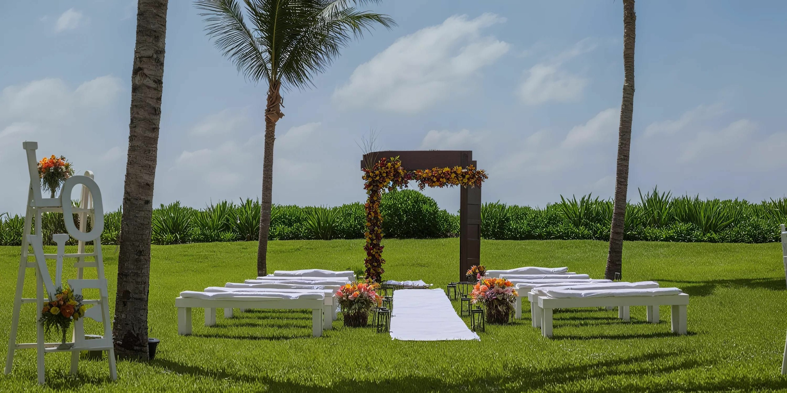 An outdoor wedding setup at Dreams Jade Riviera Cancun featuring a floral-decorated arch, benches with white covers, and a white aisle runner, flanked by palm trees on a grassy lawn under a blue sky with scattered clouds.