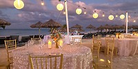 Experience a stunning beachfront setup at Hideaway at Royalton Blue Waters, featuring round tables elegantly decorated with candles and flowers. Enjoy the ambiance of string lights overhead and charming tiki huts in the background as the sun sets.