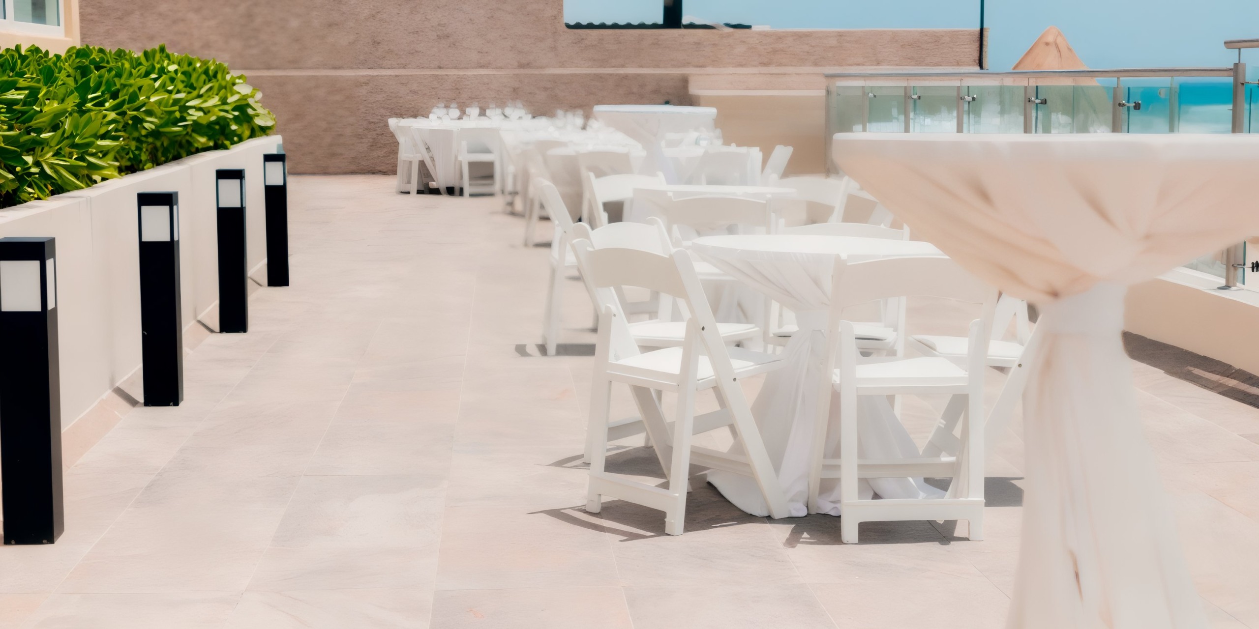 The outdoor event space at Hard Rock Cancun showcases white tables and chairs elegantly arranged on a patio, surrounded by green plants and clear barriers along the perimeter, offering a picturesque view of the sky in the background.