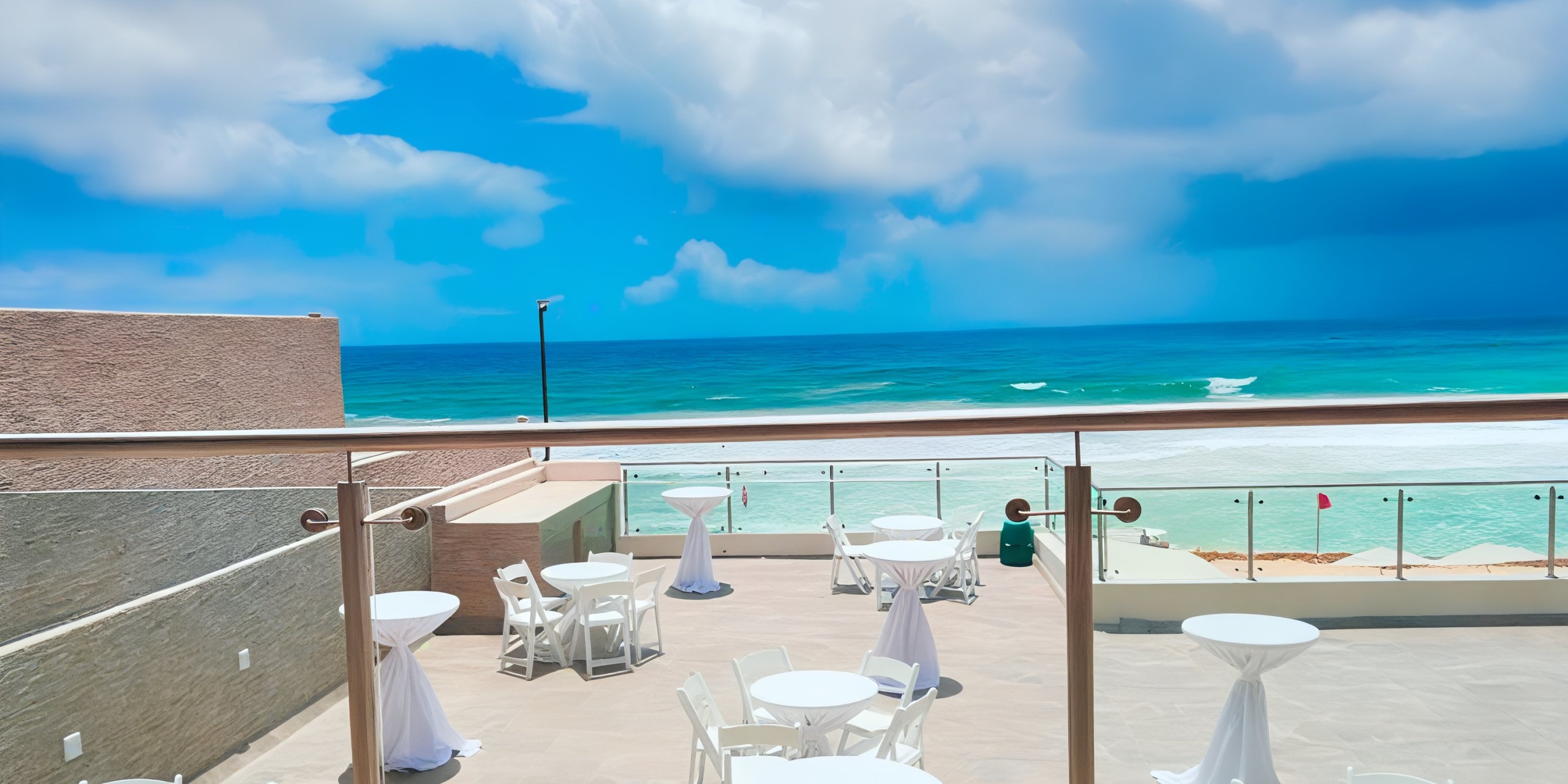 The outdoor patio at Hard Rock Cancun overlooks a beach, featuring white tables and chairs set against the stunning backdrop of the blue ocean and a cloudy sky.