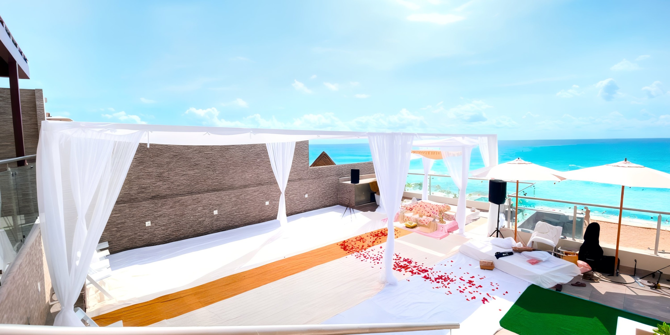 Experience an event at Hard Rock Cancun with a stunning beachfront terrace adorned in white drapery, scattered rose petals, comfortable seating arrangements, and breathtaking views of the ocean against a clear blue sky.