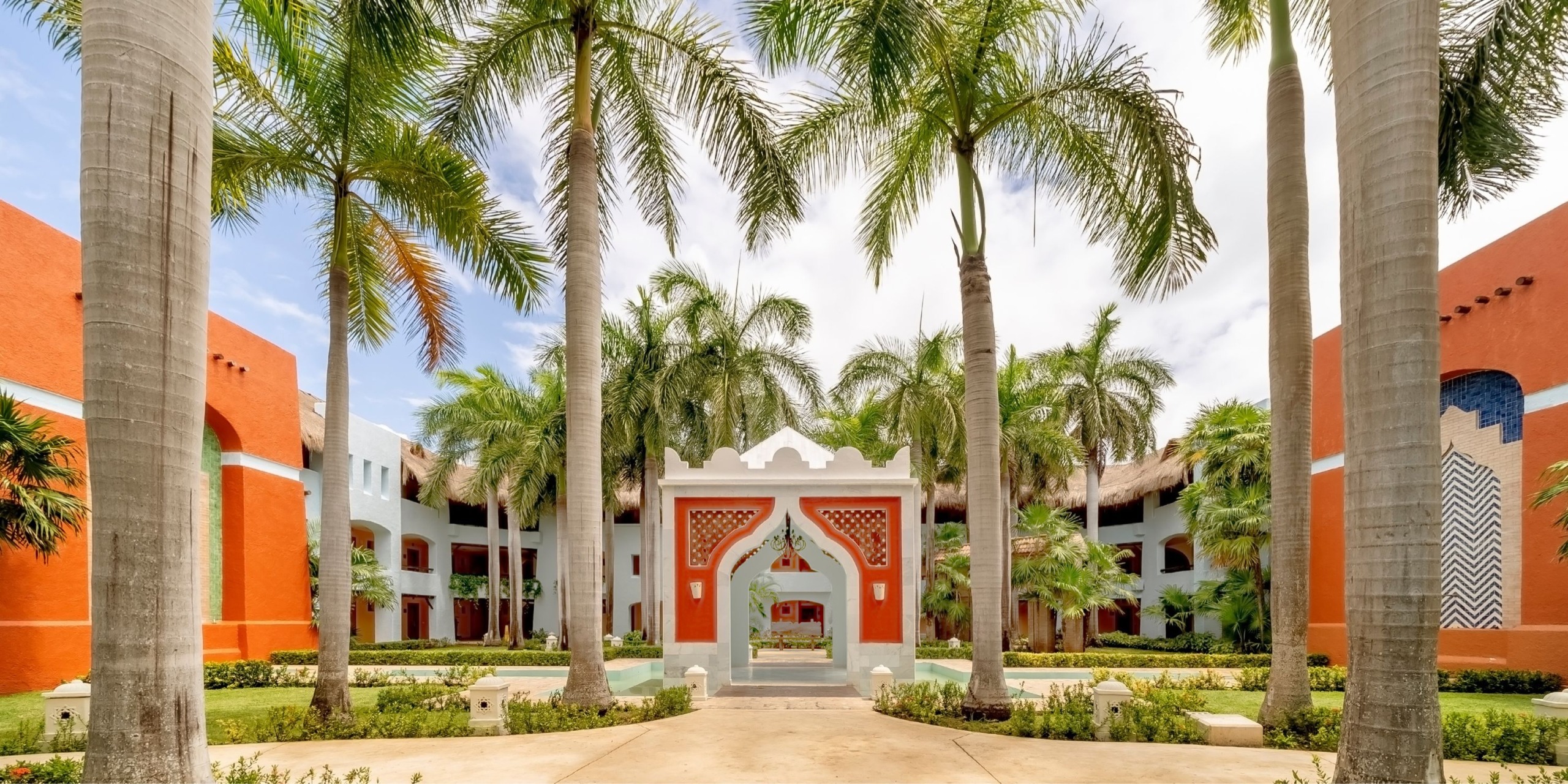 A courtyard at the Iberostar Selection Paraiso Lindo showcases tall palm trees, vibrant buildings, and a central arched structure beneath a partly cloudy sky.