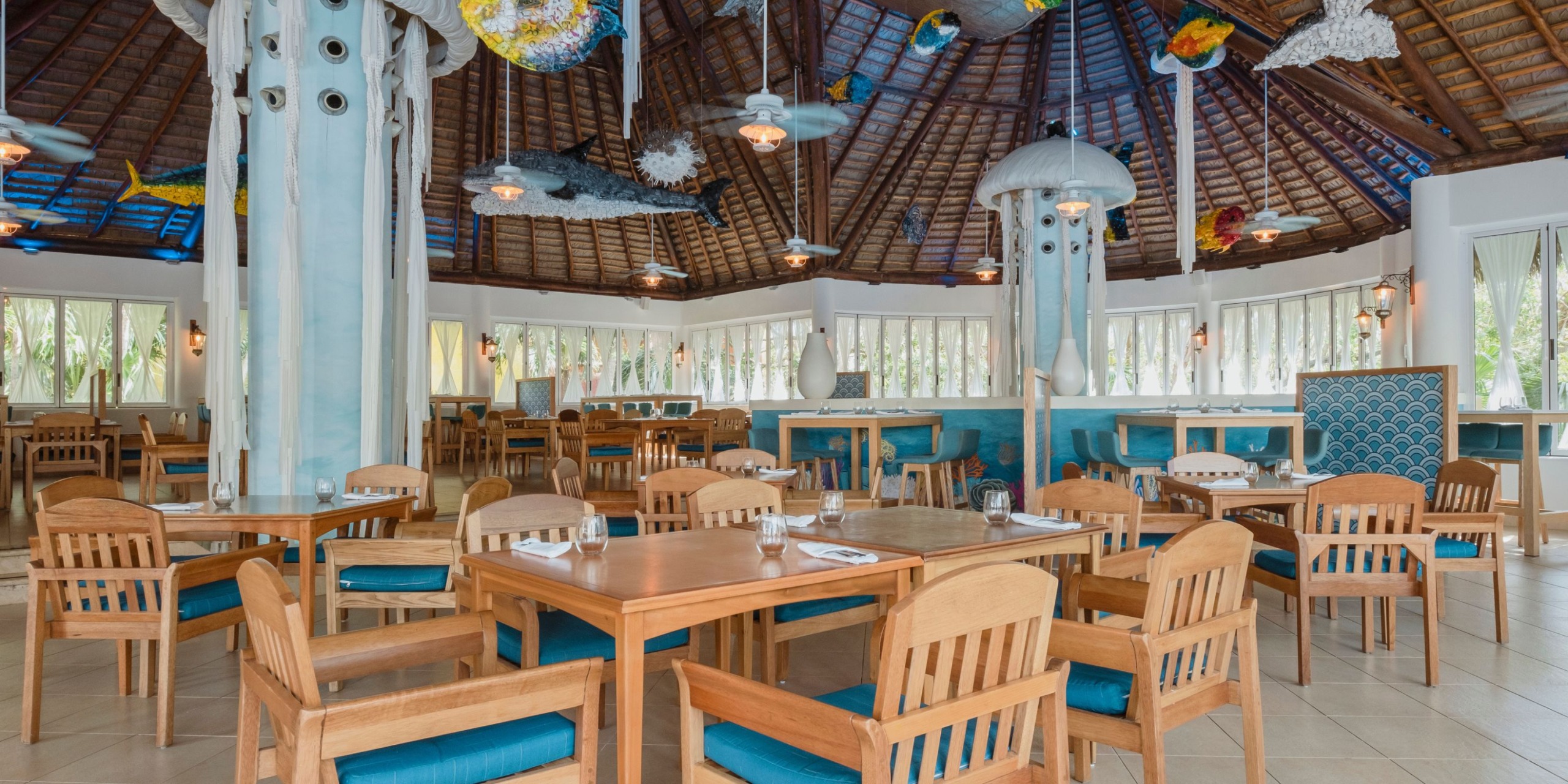 The interior of Iberostar Selection Paraiso Lindo features a spacious design with wooden tables and chairs, a high thatched ceiling, and ocean-themed decor, such as hanging fish and jellyfish sculptures, complemented by large windows.