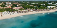 Aerial view of Iberostar Waves Paraiso Beach, a beachfront resort featuring rows of beach chairs and umbrellas, enveloped by lush greenery and buildings. Turquoise ocean waters kiss the sandy shore.
