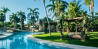 Experience a serene outdoor scene at Iberostar Waves Paraiso Beach, featuring a winding pool, tall palm trees, a wooden pergola with seating, and a thatched-roof building in the background under a clear blue sky.