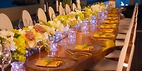 Experience the elegance of a Krystal Grand Nuevo Vallarta dining setting, featuring exquisitely arranged white chairs, a stunning floral centerpiece, crystal-clear wine glasses, and gilded cutlery.