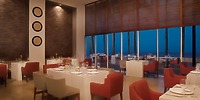 The Krystal Grand Nuevo Vallarta offers a contemporary dining experience with red and beige chairs, white tablecloths, and decorative wall plates. Large windows showcase a blue evening sky.