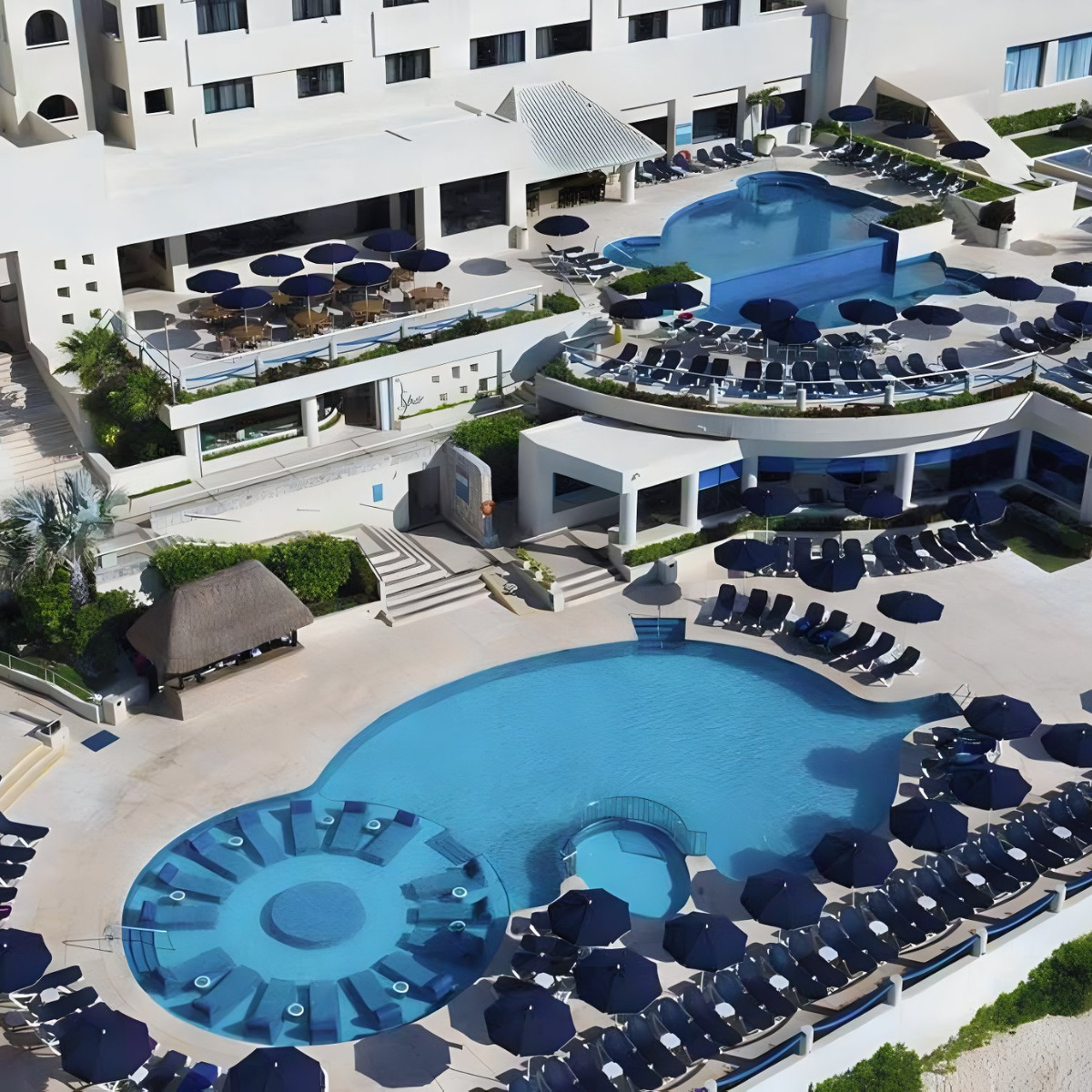 Aerial view of the Occidental Tucancun, a popular destination for weddings, with a large swimming pool surrounded by loungers and parasols, adjacent to a white building complex.