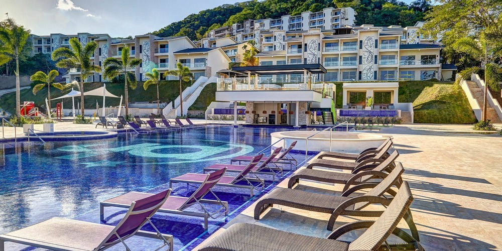 A pool area with lounge chairs is situated in front of the modern white buildings of Planet Hollywood Costa Rica on a sunny day, featuring palm trees and a hillside in the background.