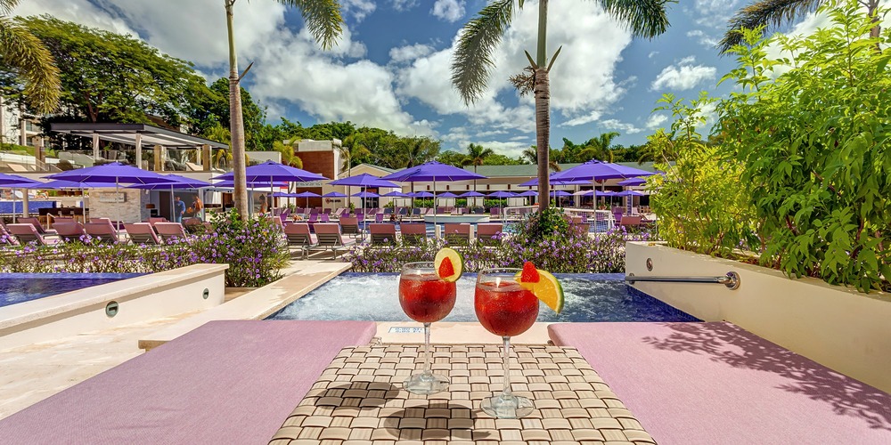 Two Planet Hollywood Costa Rica glasses of red drinks on a woven mat by a pool, with purple umbrellas and lounge chairs in the background under a partly cloudy sky.