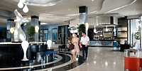 A couple strolls through the contemporary lobby of Hotel Riu Palace Costa Rica, featuring abstract sculptures, sweeping design elements, and a reception desk in the background.
