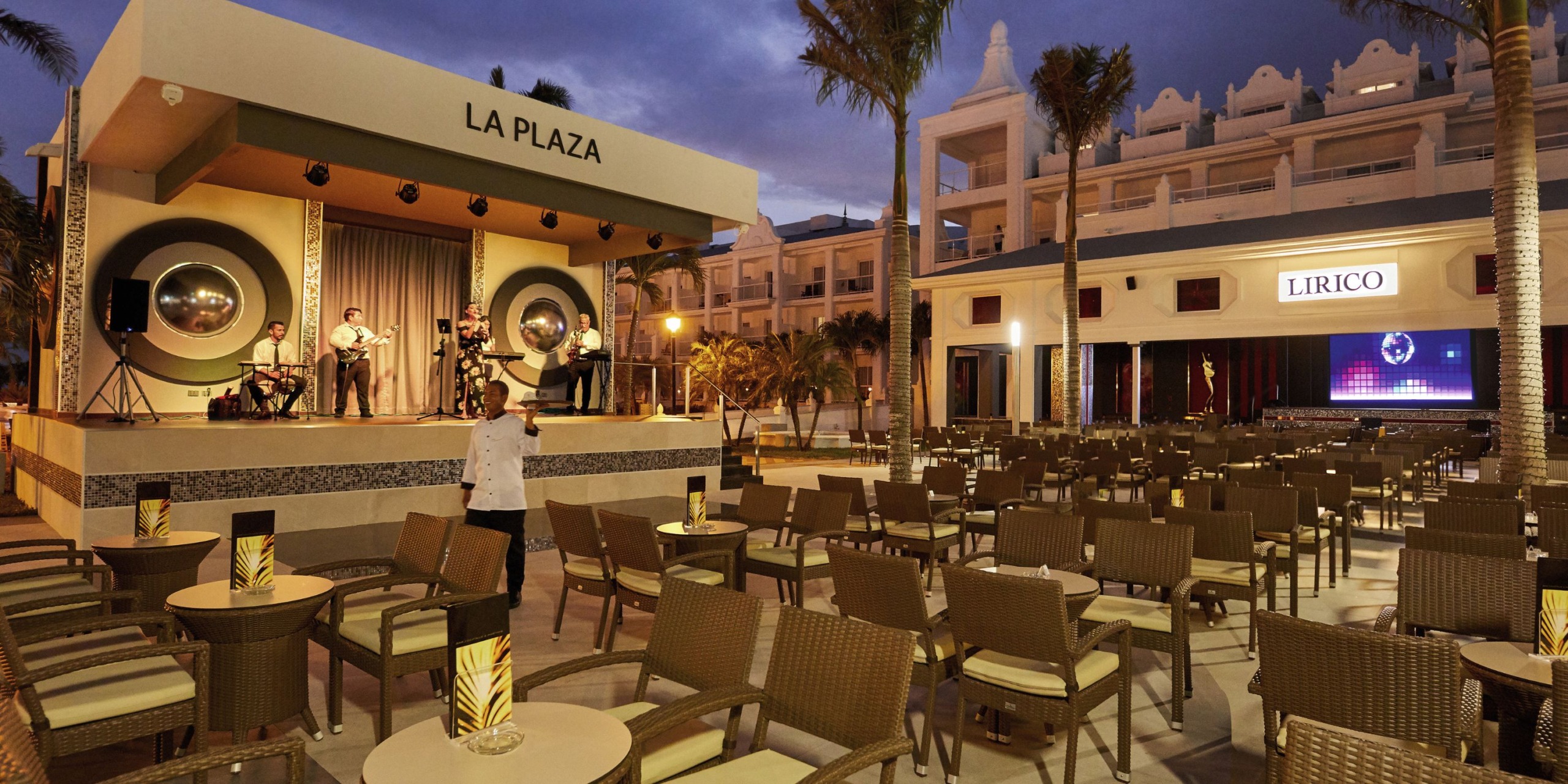 Lively outdoor dining area at Hotel Riu Palace Riviera Maya, complete with wicker chairs and tables set before a stage labeled "La Plaza," where musicians perform. In the background, a building labeled "Lirico" stands under the twilight sky.