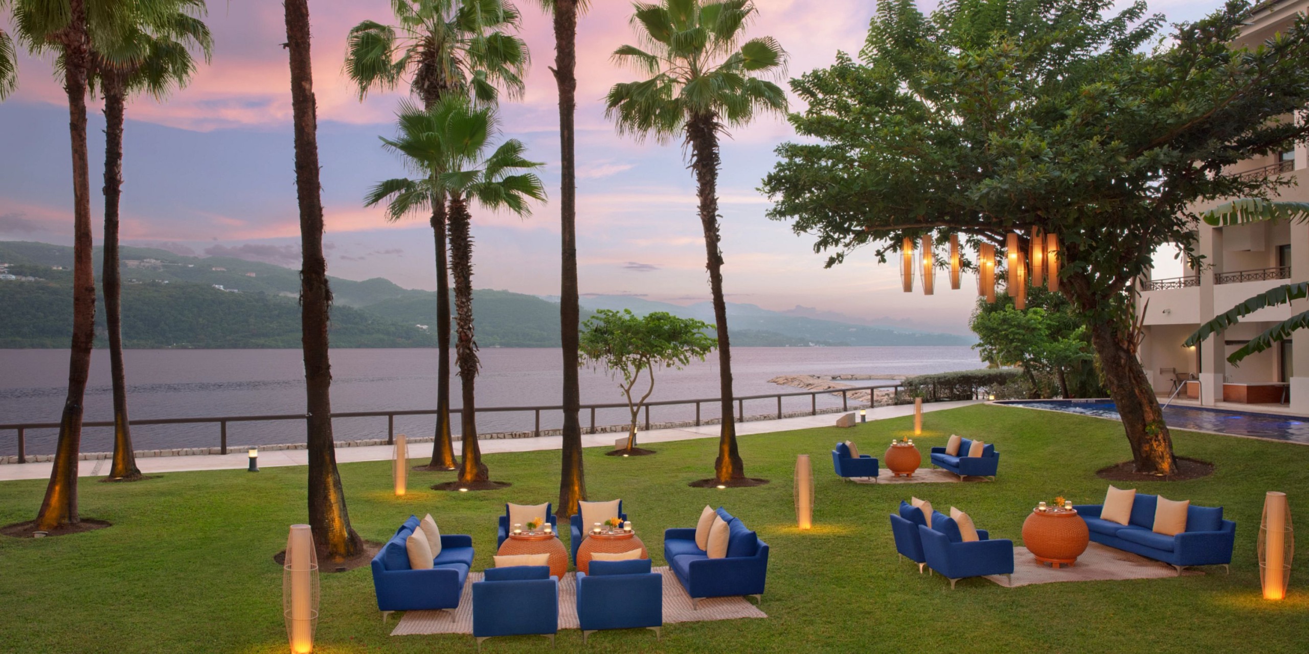 A charming seating area at Secrets St. James Montego Bay features blue chairs and tables on a lush grassy lawn. Surrounded by tall palm trees and decorative lights, this peaceful spot overlooks a shimmering body of water with distant hills under a pink sky.