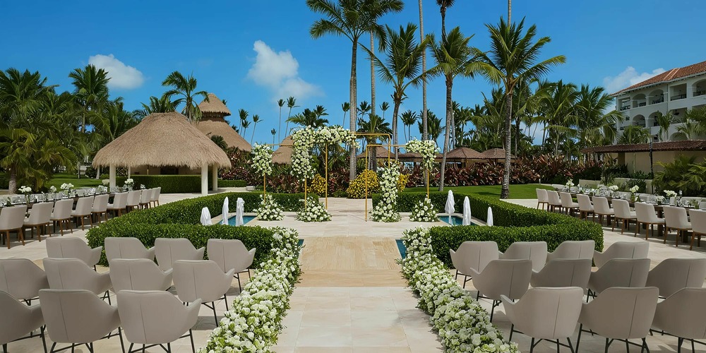 Experience an outdoor wedding ceremony at Secrets Royal Beach, featuring white chairs arranged in rows, a flower-lined aisle, and a decorative arch surrounded by lush greenery and palm trees under a clear blue sky.