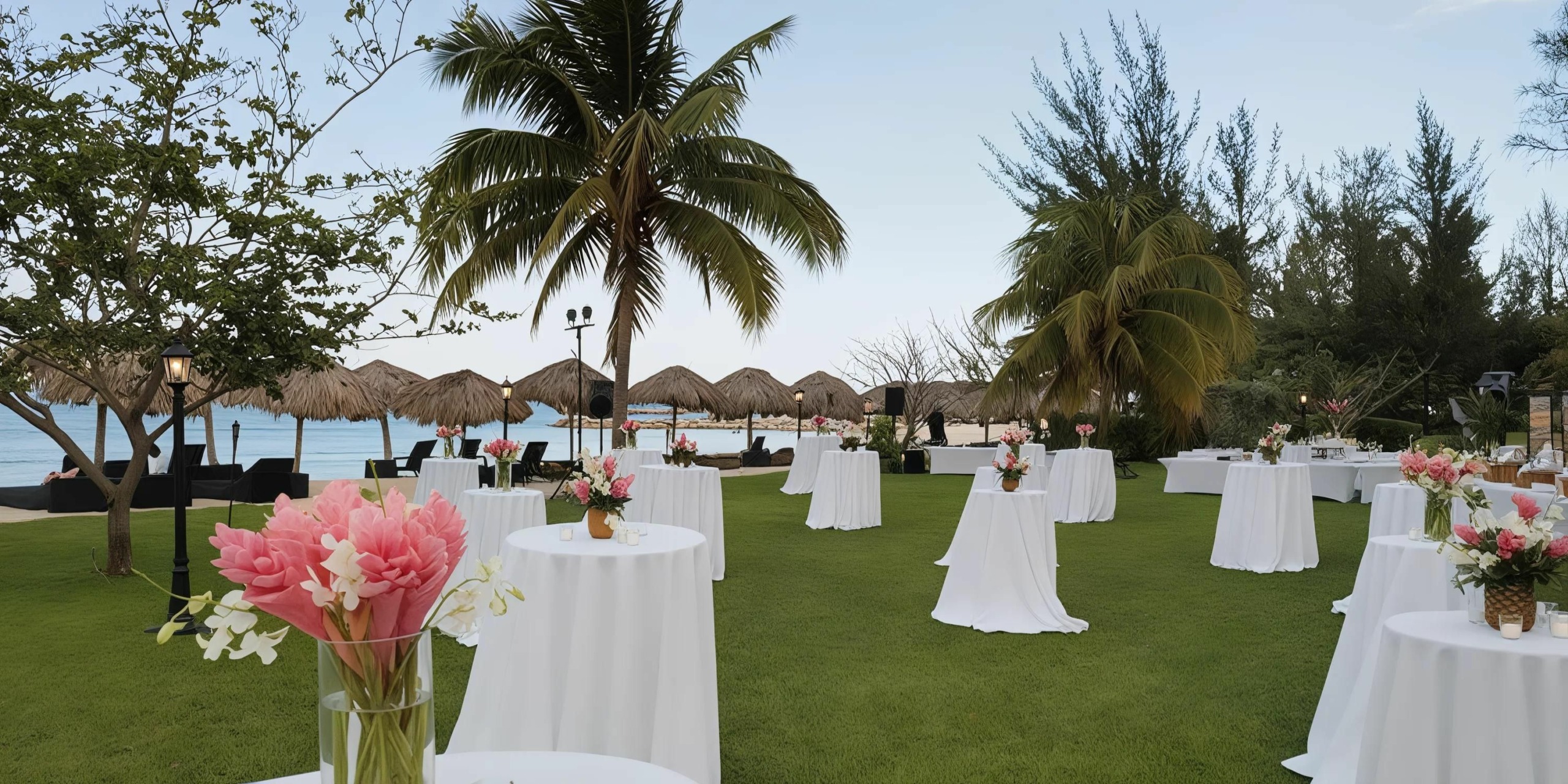 Outdoor event setup on a grassy lawn at Secrets St. James Montego Bay with round tables covered in white cloth, pink floral arrangements, palm trees, and thatched-roof shelters in the background near a body of water.