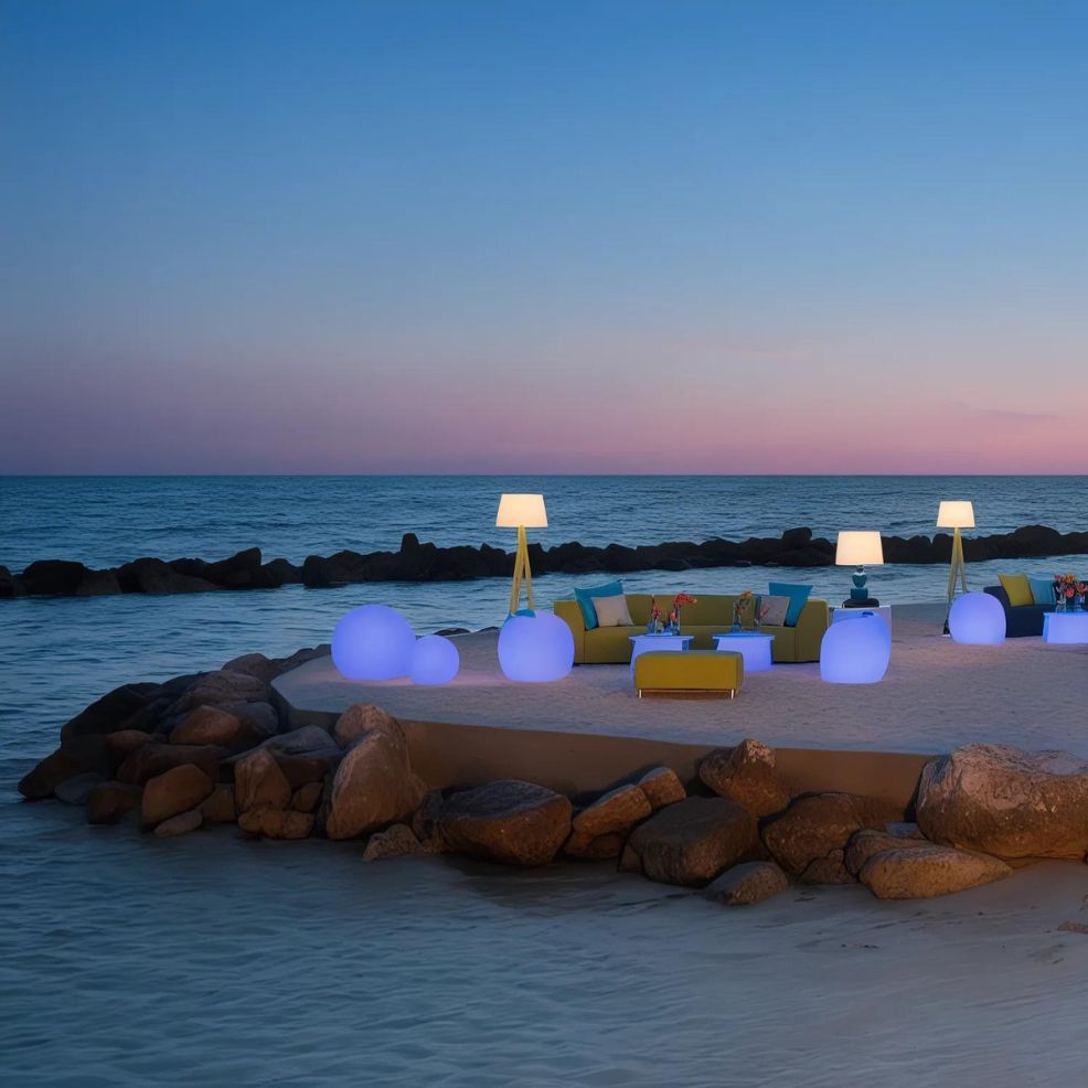 Outdoor lounge area with illuminated lamps and seating on a sandy beach, bordered by rocks, during dusk. The sea and sky show soft hues of blue and pink in the background.