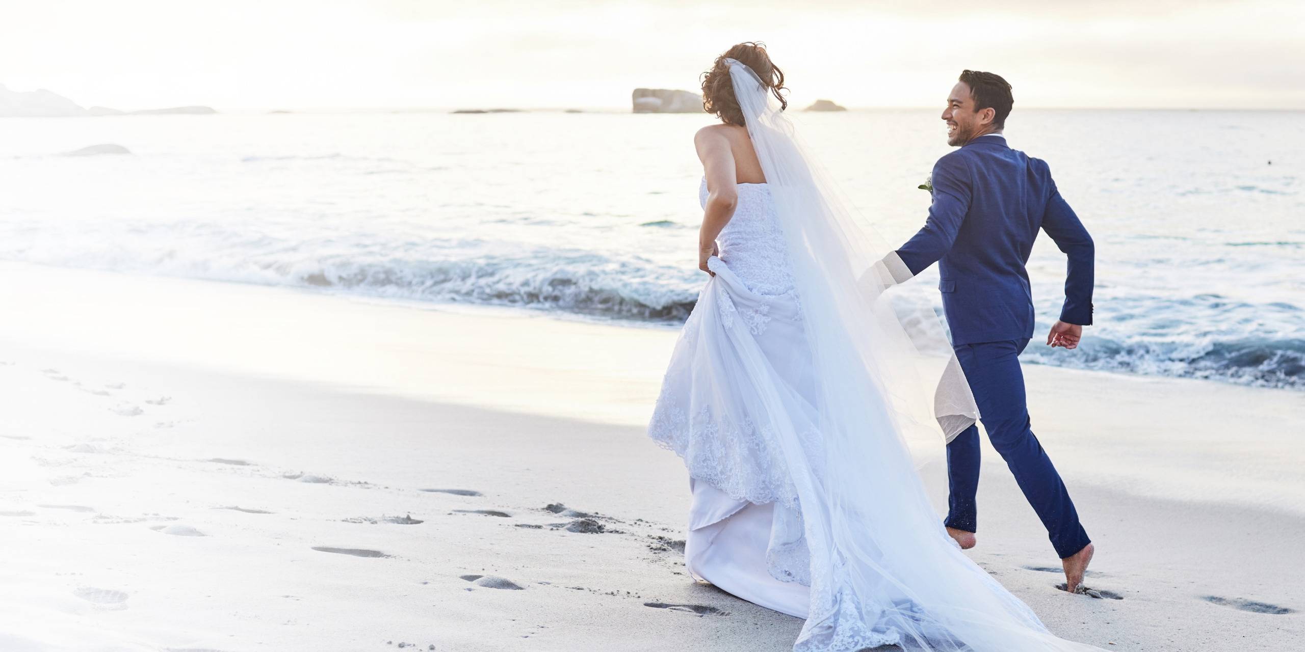 Husband and wide, groom and bride in white bridal dress run along an ocean beach during their destify destination wedding