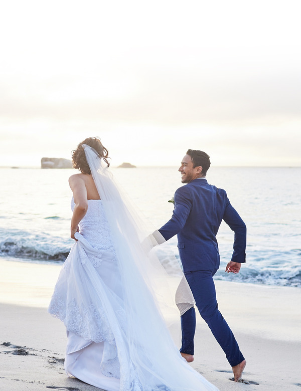 Husband and wide, groom and bride in white bridal dress run along an ocean beach during their destify destination wedding