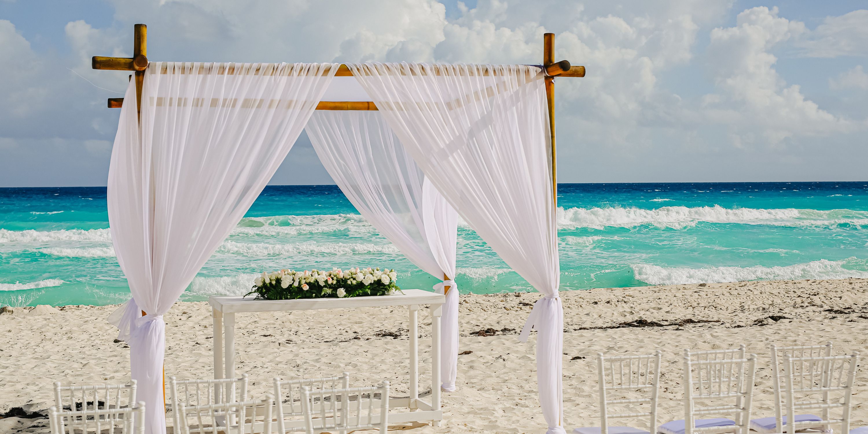 Destination wedding altar at Beach Palace Cancun with white drapes, ocean backdrop, and chairs on sand.