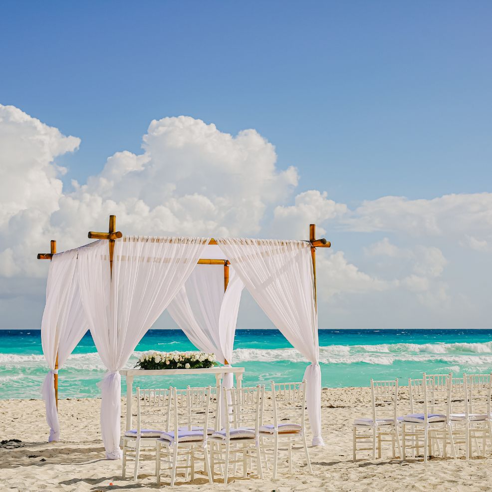 A destination wedding setup with a white-draped arch and chairs on a sandy beach overlooking the ocean.