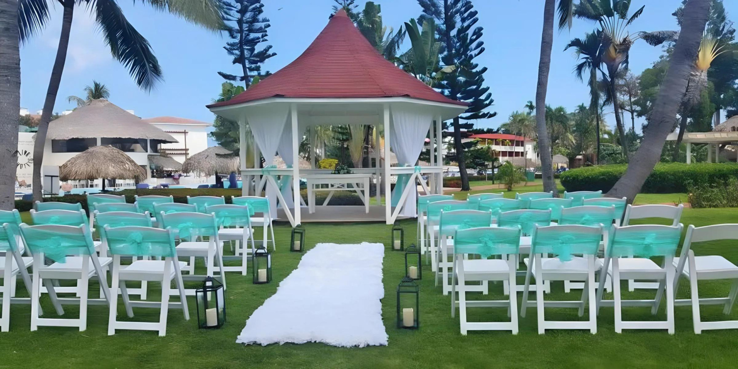 Destination outdoor wedding setup with white chairs and a gazezebo, surrounded by palm trees and green grass, under a clear sky.