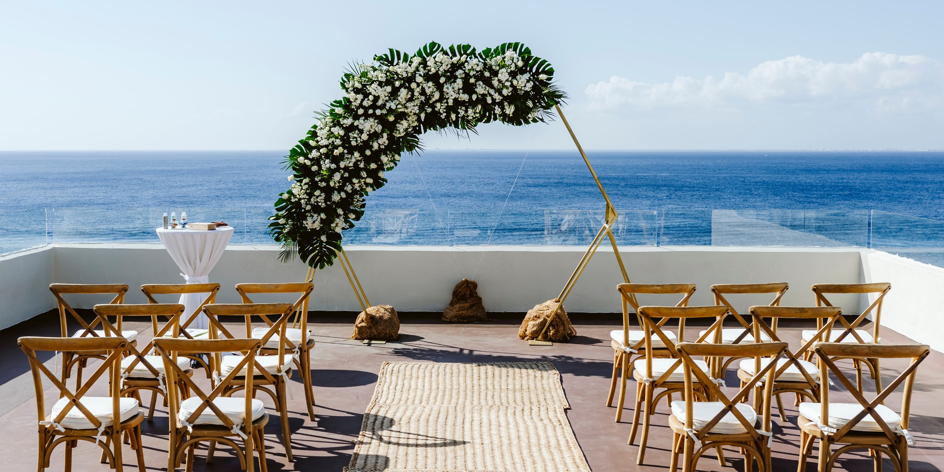 Destination wedding at Cozumel Palace with wooden chairs and a floral arch overlooking the sunny ocean.