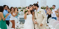 The wedding couple walks the aisle at a destination wedding as guests cheer and toss flower petals.