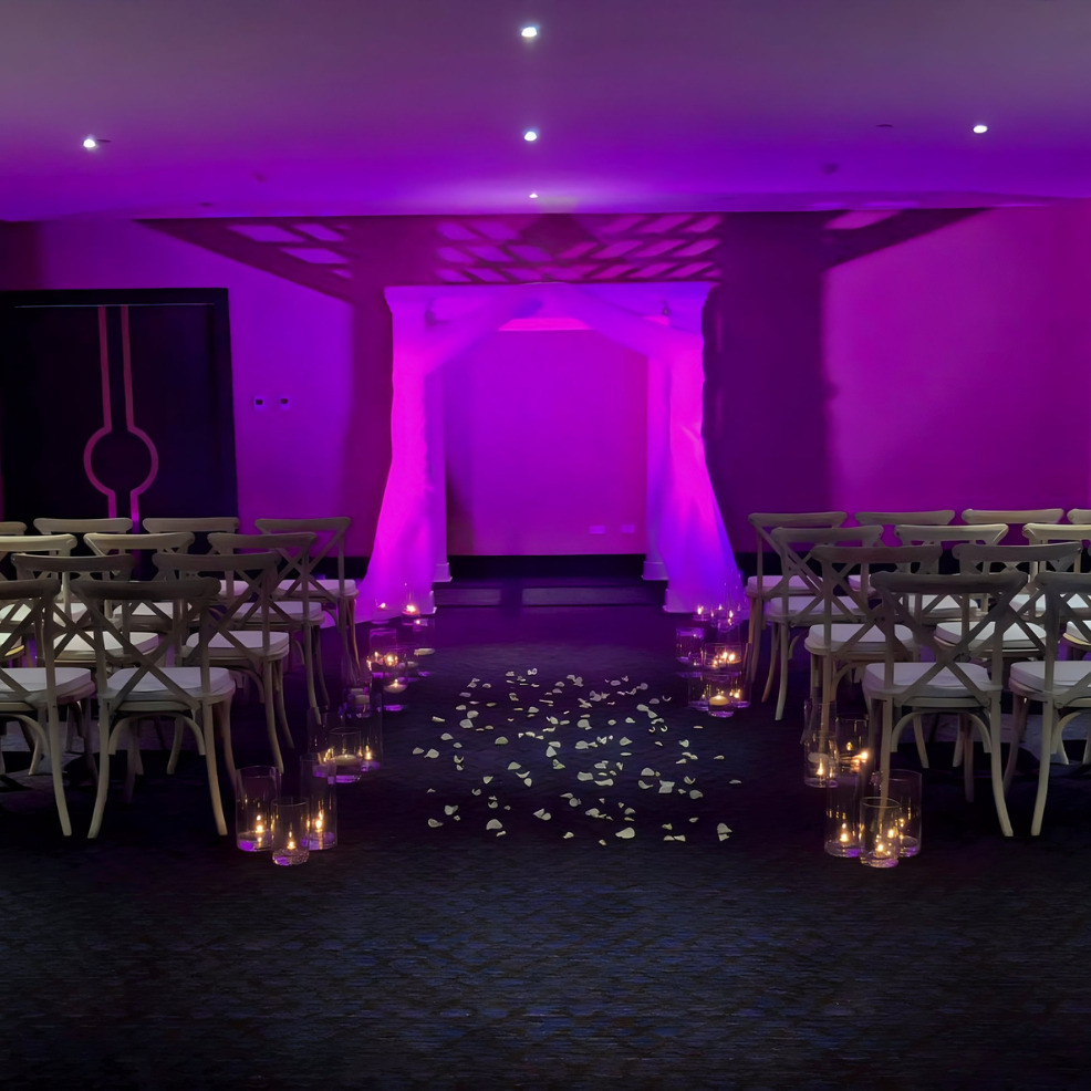 Wedding aisle decorated with white rose petals and lanterns, illuminated by purple lighting under a modern arched structure in a destination wedding venue.