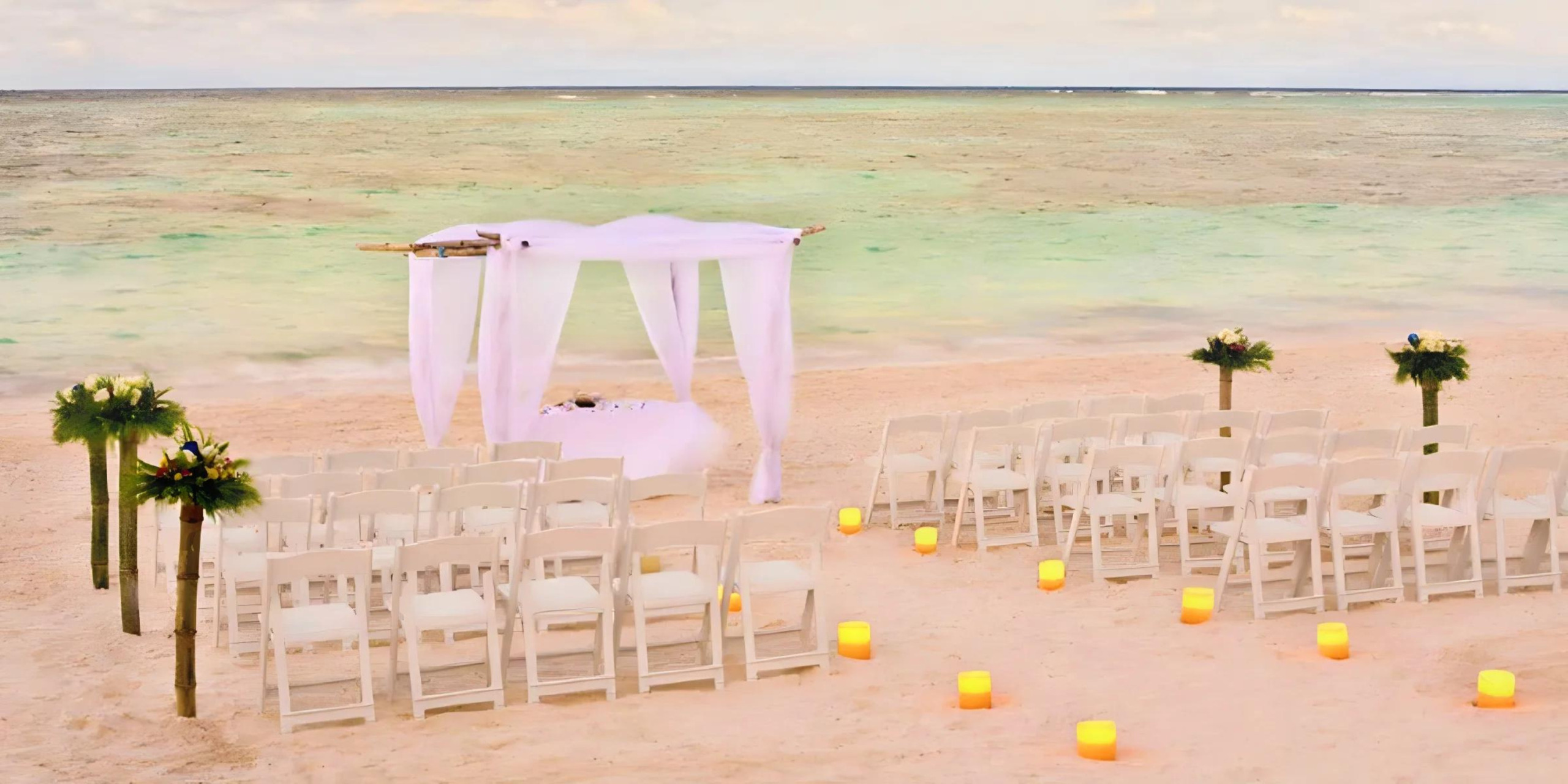 Destination beach wedding setup with white chairs, a canopy, and candles on the sand at sunset.