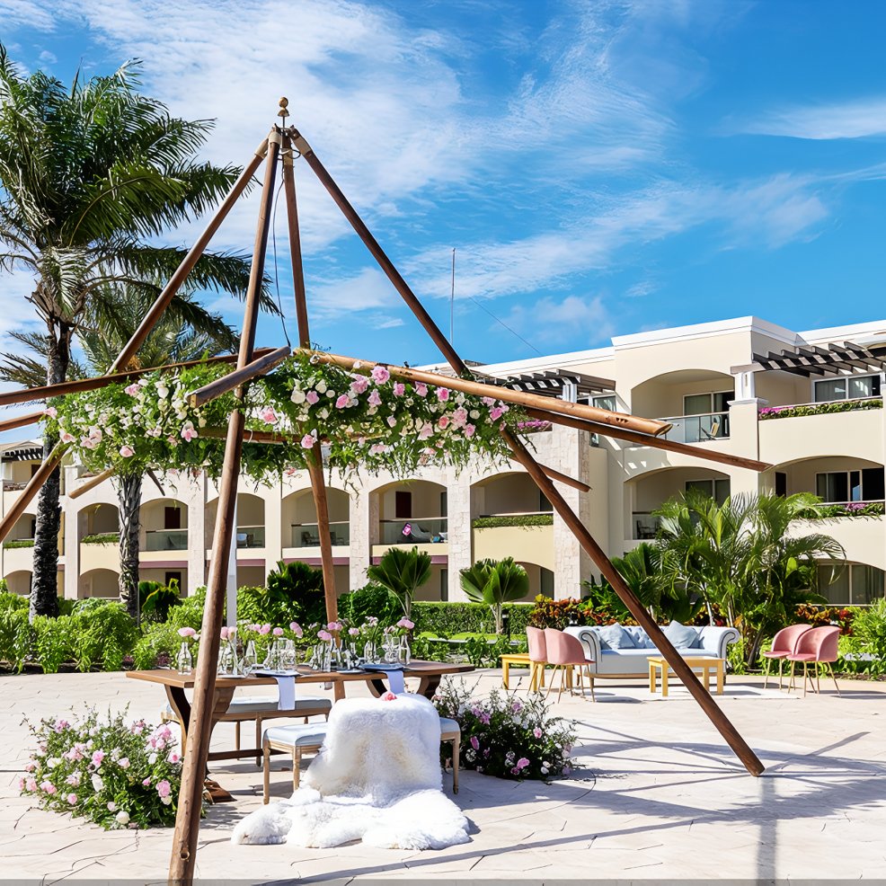 Destination wedding table set beneath a floral arch outside a resort building on a bright, sunny day.