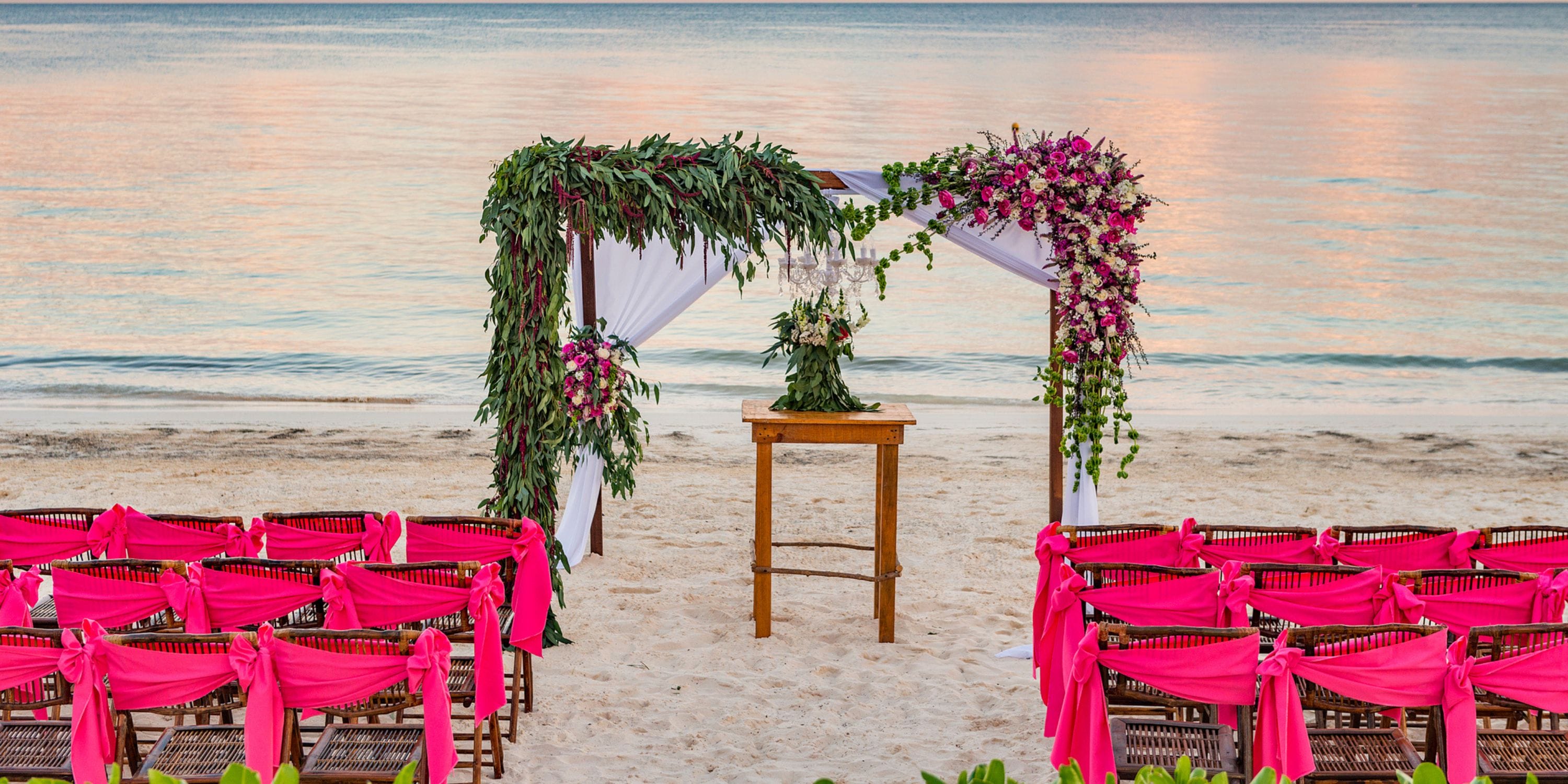 A destination wedding altar with pink chairs is arranged on the sandy beach at Ocean Coral and Turquesa.