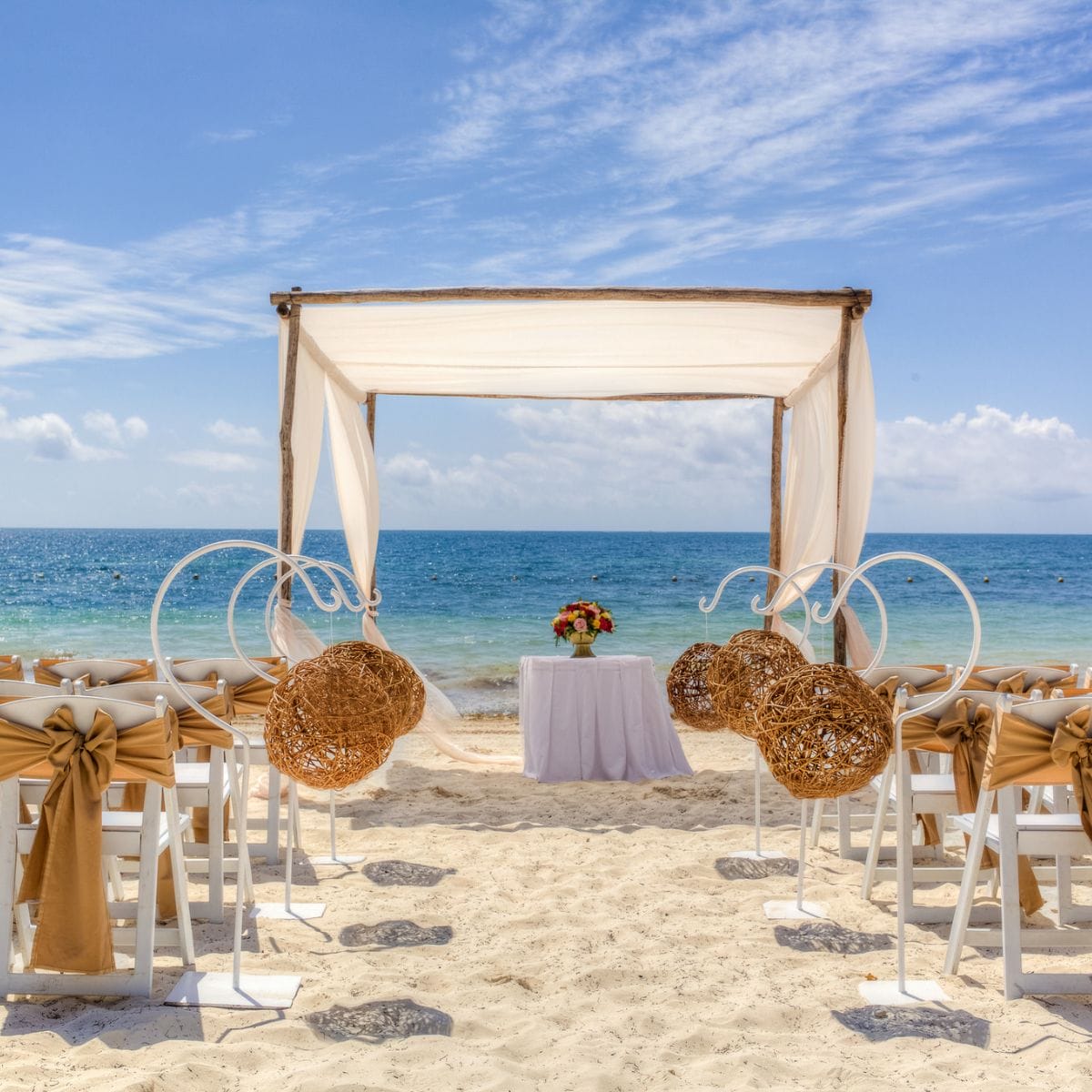 Destination wedding setup on a beach with chairs, aisle, and canopy arranged toward the ocean under sunny skies.