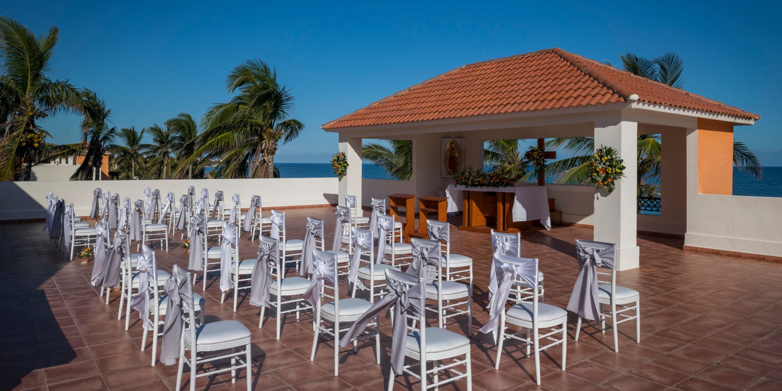 Rows of white chairs arranged for a destination wedding on a tiled terrace overlooking palm trees and ocean.
