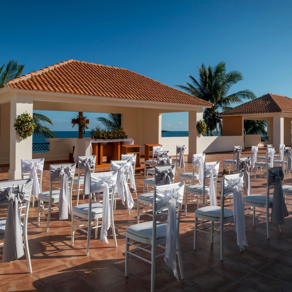 White chairs with bows arranged for a destination wedding ceremony by the ocean under a blue sky.