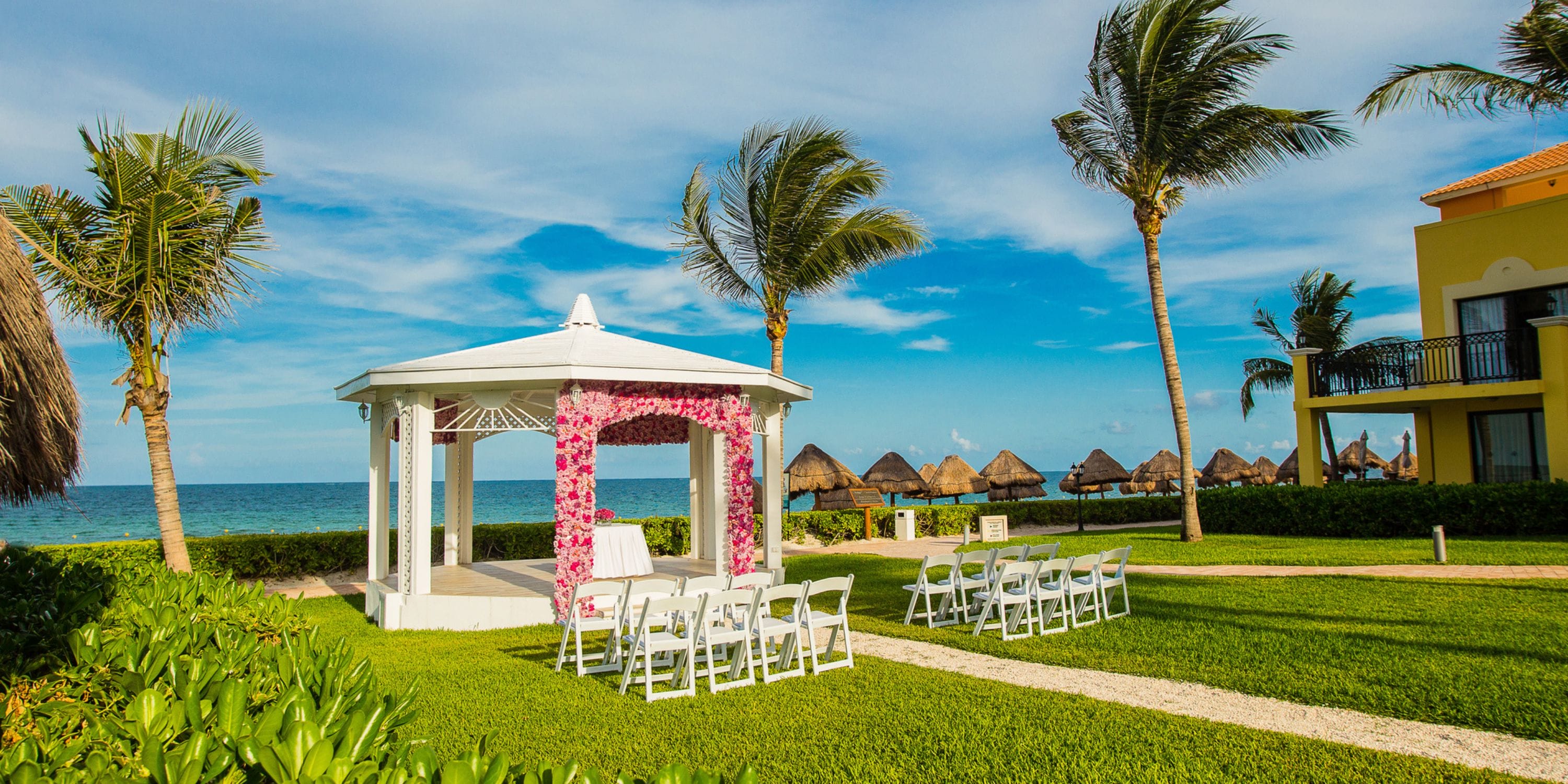 Ocean Coral and Turquesa: White gazebo set for a destination wedding with pink flowers by the sunny ocean.