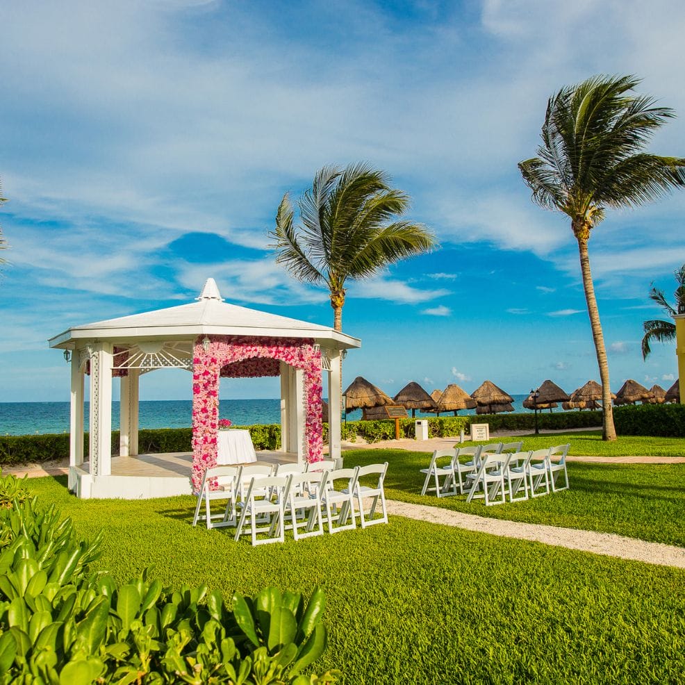 Destination wedding setup with a white gazebo, pink floral arch, white chairs, and palm trees by the beach.