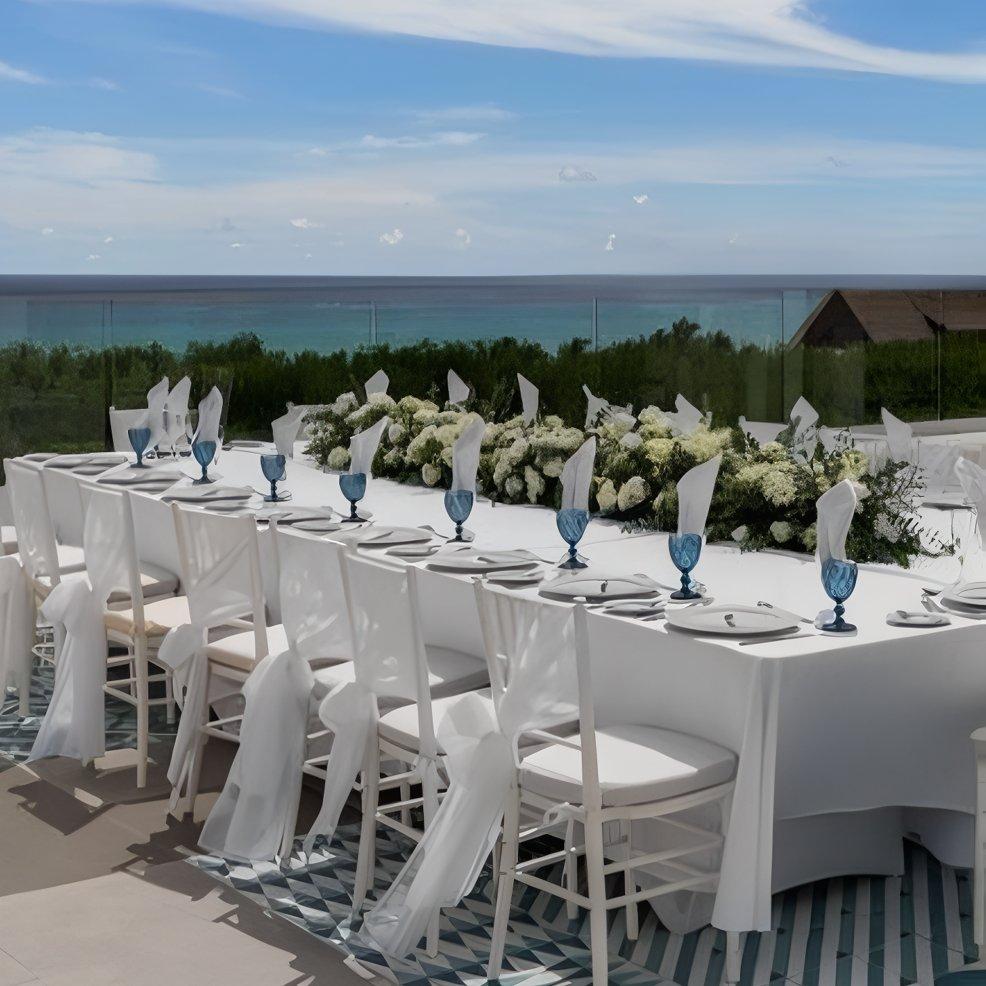 Elegant destination wedding table with white linens, blue glasses, and floral centerpiece by the ocean.