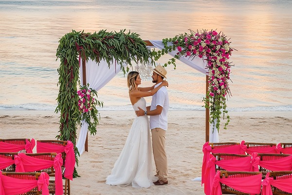A couple embraces beneath a floral arch at their destination wedding at Ocean Coral and Turquesa, surrounded by pink seating on the beach.