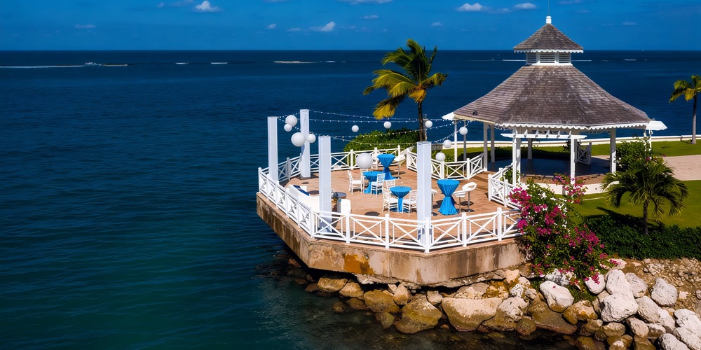Moon Palace Jamaica: A picturesque gazebo on the coastal deck provides a stunning ocean view, surrounded by palm trees and ample seating, making it an ideal setting for a destination wedding.